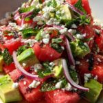 Een grote witte schaal met gegrilde watermeloensalade, met duidelijk zichtbare stukjes feta, avocado, rode ui en verse munt.