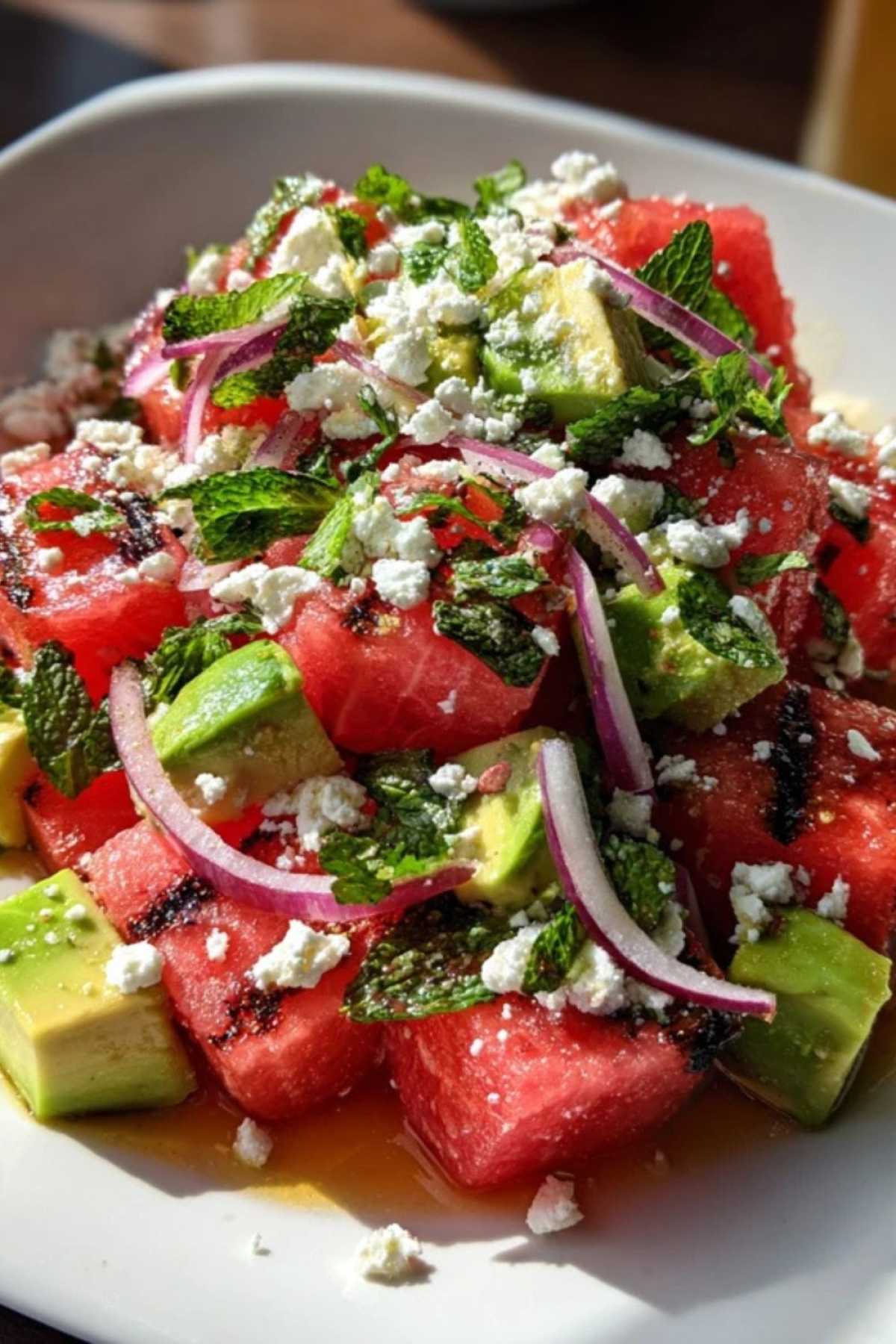 Een grote witte schaal met gegrilde watermeloensalade, met duidelijk zichtbare stukjes feta, avocado, rode ui en verse munt.