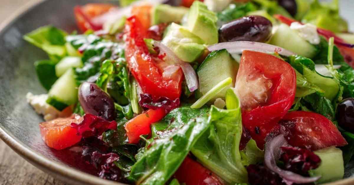 Een hand giet een goudbruine balsamico-honingdressing uit een glazen potje over een kom met gehakte salade.