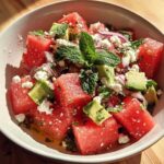 Verse ingrediënten voor een gelaagde salade, waaronder gesneden komkommer, paprika, tomaat en feta.