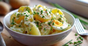 Cremiger deutscher Kartoffelsalat in einer weißen Schüssel, garniert mit frischem Schnittlauch.